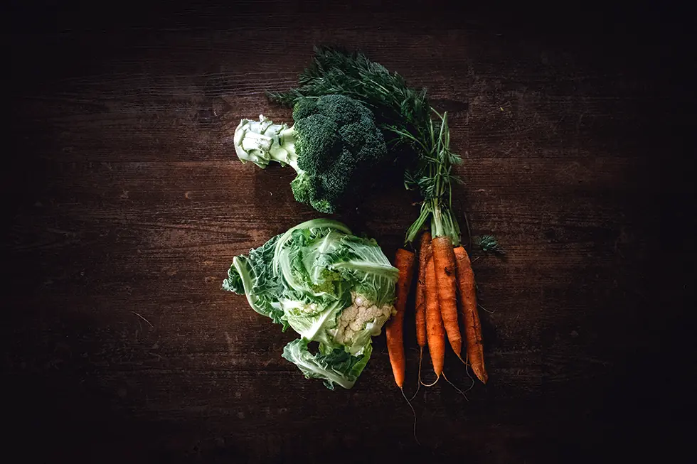 Broccoli, a bunch of carrots with their tops still on, and a cauliflower wrapped in its own leaves, laid out on a dark wooden table in low light.